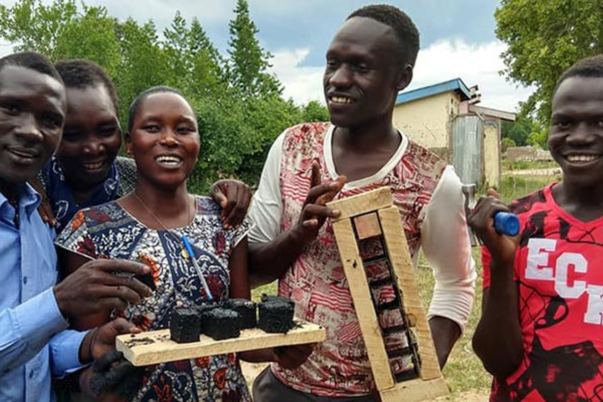 A group of smiling individuals outdoors holding handmade tools and materials, showcasing a participatory design project.